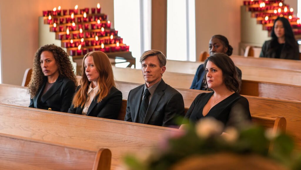 Cena da terceira temporada de Yellowjackets. Da direita para a esquerda, Tai (Tawny Cypress), Van (Lauren Ambrose), Jeff (Warren Kole) e Shauna (Melanie Lynskey) se sentam no banco de uma igreja bem iluminada, acompanhando o velório de alguém. Tai é uma mulher negra de cabelos encaracolados e longos, usando um paletó escuro com botões dourados. Van é uma mulher caucasiana de cabelos ruivos e longos com um paletó escuro por cima de uma camisa branca abotoada. Jeff é um homem caucasiano de cabelos loiros e curtos, usando um paletó escuro e uma gravata verde escura. Shauna é uma mulher caucasiana de cabelos negros, usando um vestido preto com uma corrente dourada no pescoço. A igreja é bem iluminada, com velas vermelhas acesas ao lado das janelas abertas. Atrás do grupo, duas outras mulheres se sentam sozinhas em bancos mais afastados.