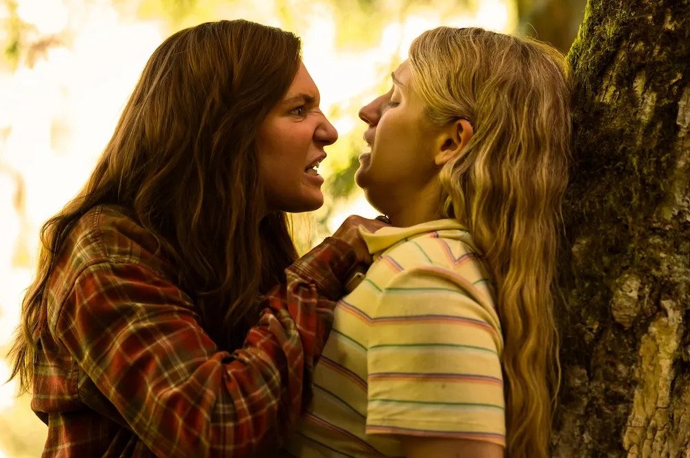 Cena da terceira temporada de Yellowjackets. Shauna (Sophie Nélisse) ameaça Melissa (Jenna Burgess) com uma faca, pressionando-a contra o tronco de uma árvore. Shauna é uma garota caucasiana de cabelos castanhos e longos, usando uma camisa xadrez vermelha de mangas longas, segurando uma faca com a mão direita, pressionando-a contra o pescoço da outra. Melissa é uma garota caucasiana de cabelos loiros e longos, usando uma camiseta de mangas curtas clara e listrada horizontalmente, se inclinando para longe de Shauna, que a olha com uma expressão irada. Atrás delas, o fundo de uma floresta bem iluminada e fora de foco.