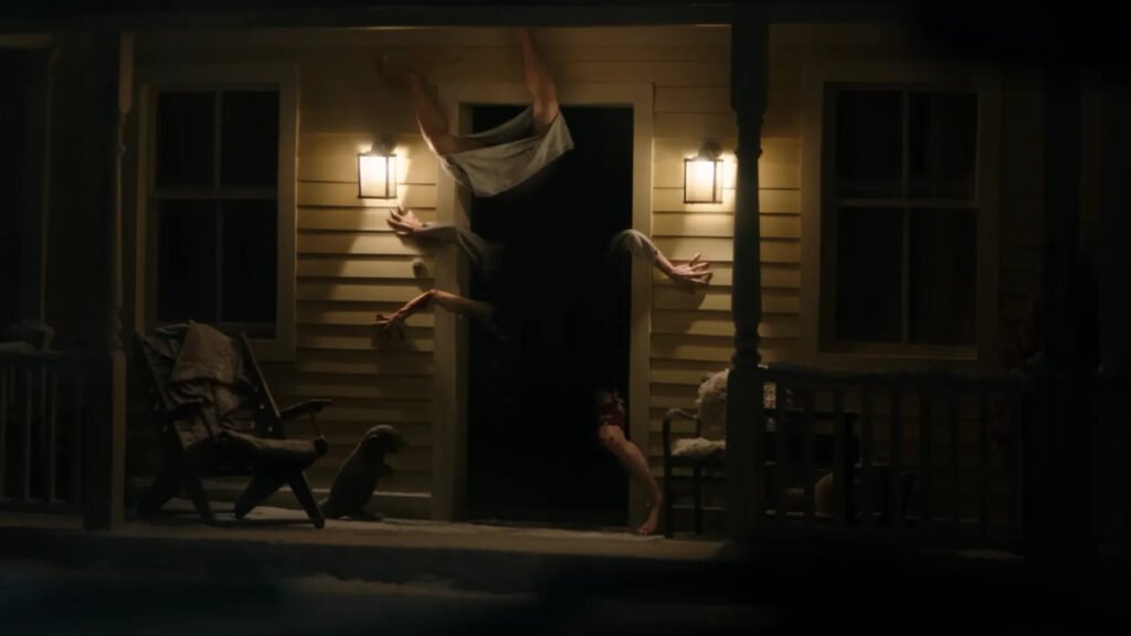 Cena do filme Resident Evil. Três mãos e três pernas saem da porta escurecida de uma casa, iluminada em ambos os lados por lanternas amarelas nas paredes. Um par de pernas surge na parte de cima, virados para o teto, enquanto a outra perna aparece na parte direita. Dois braços agarram a parede esquerda e uma agarra a direita. Há janelas em ambas as paredes da casa e duas cadeiras no pórtico.