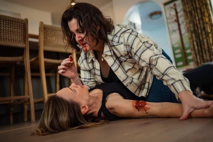 Cena da terceira temporada de Yellowjackets. Shauna (Melanie Lynskey) está sobre Melissa (Hilary Swank), forçando-a a comer um pedaço de sua própria carne, que ela mordeu de seu braço direito. Shauna é uma mulher caucasiana de cabelos escuros, usando uma camisa xadrez clara sobre uma camiseta escura e calças jeans. Melissa é uma mulher caucasiana de cabelos loiros e longos, usando uma regata preta. Atrás delas, uma mesa de jantar com três cadeiras de madeira dispostas ao lado e, à direita, um arco que dá para uma sala de estar.